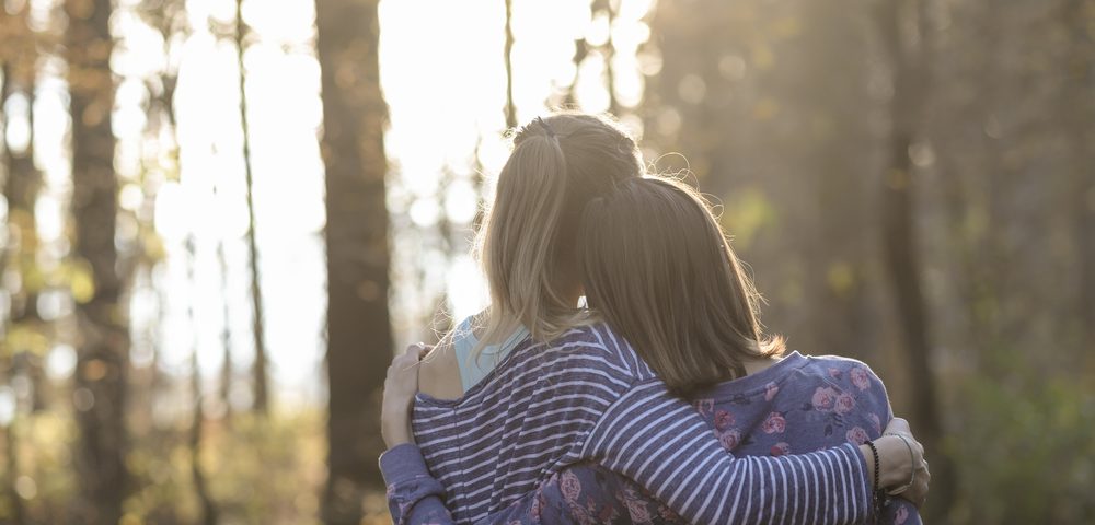 Cystic Fibrosis News Today \ A stock image of two women - either sisters or friends - hugging in a forest, with bright, out-of-focus sunlight piercing the trees in the background