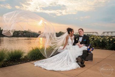 Cystic Fibrosis News Today | A professional photograph captures Jared and Nicole on their wedding day. They are seated on a bench overlooking a river at sunset. Nicole has her legs flung over Jared's lap, and her veil billows out behind her.