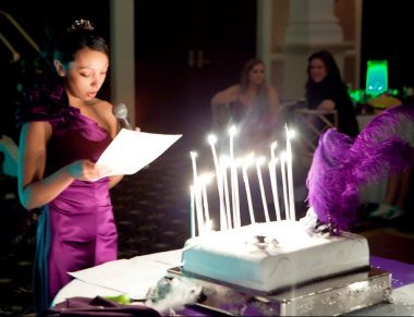 Make-A-Wish | Cystic Fibrosis News Today | Nicole wears a purple dress to celebrate her 18th birthday. She is holding a microphone and reading from a white paper in her hands in front of a cake with many candles. Two women are seen at a table in the background.