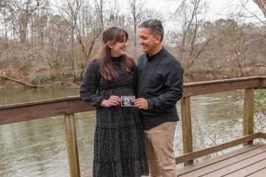 CF and infertility | Cystic Fibrosis News Today | Joanie and Jorge Santander stand on a deck overlooking a river in a wooded area and hold a photo of an ultrasound. 