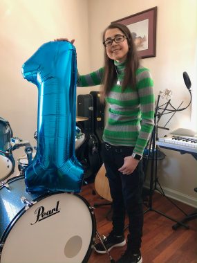 CF community | Cystic Fibrosis News Today | Dressed in a green and gray striped sweater, Heather McCoy holds up a blue birthday ballon in the shape of the number one. It sits on a blue drum set..