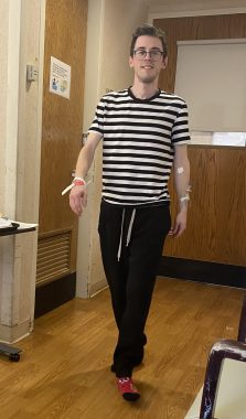 A young man in a black-and-white-striped shirt paces in a hospital room. He's wearing black sweatpants and has hospital bracelets on his wrist. He wears glasses and is smiling for the camera. The photo is meant to demonstrate the cramped space of a hospital room, and how hard it is to get any exercise by walking around. 