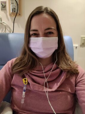 A close-up selfie of a woman in an infusion chair at a hospital or clinic. She's wearing a mask and headphones.
