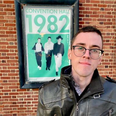 A young man wearing a leather jacket and glasses poses in front of a brick wall featuring a poster of the Clash. He's carrying a machine to infuse antibiotics through an IV and PICC line, but it's just out of the frame.