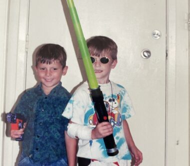 Two young boys play "Star Wars"; one holds a gun and wears a smile, while the other, wearing round sunglasses, holds a green light saber. 