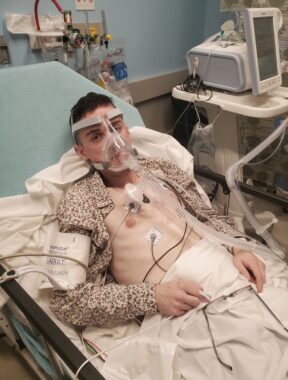 A young man lies at an incline on a hospital bed. He's wearing a cheetah-print collared shirt that is unbuttoned and pushed to either side, allowing several monitors to be attached to his chest. He's also wearing a large CPAP machine on his head that prohibits him from wearing a face mask. Medical equipment is visible behind him.