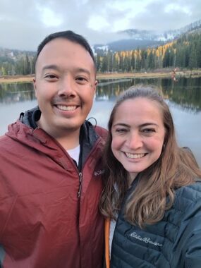 Lara and Christopher stand in front of a lake, both smiling and wearing jackets. It appears to be fall, as the mountains in the background are partially covered in snow, and a section of the foliage has turned gold. 