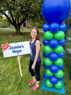 A woman in a blue tank top and black leggings stands in a green grass with a large tree in the background. She holds a sign with the word "hope" and has her back to a column of blue and green balls.