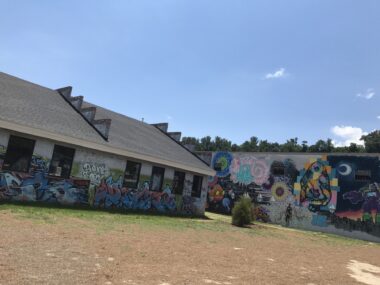 Two buildings are covered in vibrant graffiti amid a mostly brown dirt field, with a little green grass near each building.