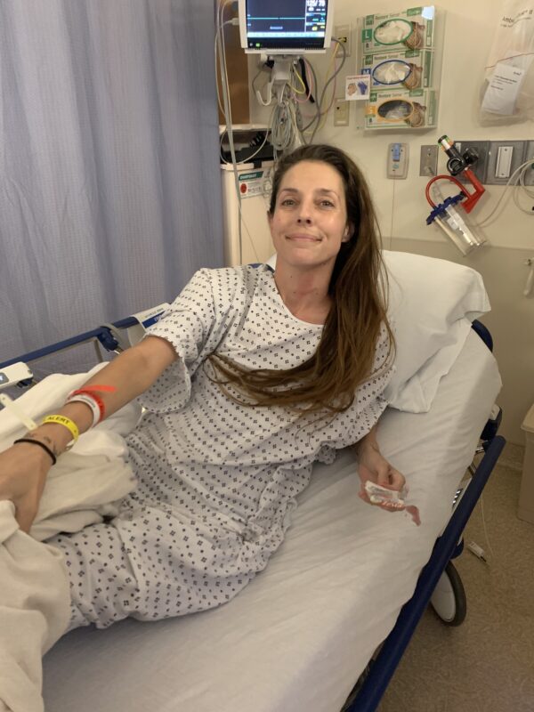 A woma n with long brown hair is in a hospital gown while lying in a hospital bed. Multicolored straps are on her right arm.