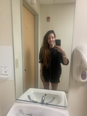 a young woman with long brown hair and a black top and shorts stands before a bathroom sink, taking a selfie with the mirror above it.