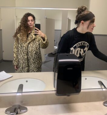 Two women are seen in a bathroom mirror. One, holding a phone camera, wears a long coat with leopard prints and has long brown hair. The other has her brown hair tied in a bun and wears what appears to be a long-sleeve black T-shirt with an AC/DC band logo.