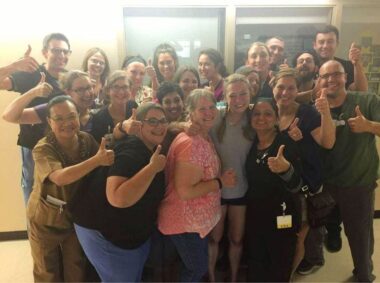 A young woman in a gray T-shirt and short smiles for a photo surrounded by her mom, sister, and nearly 20 nurses. They appear to be standing in a hospital room, and most of the nurses are giving a thumbs-up.