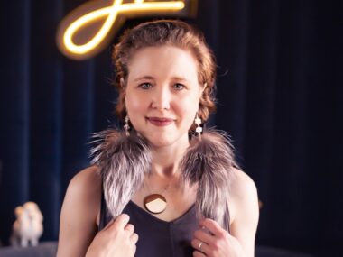 A colorful and well-composed headshot of a woman with an intriguing outfit and large, fluffy, feathered earrings, set to a black background.