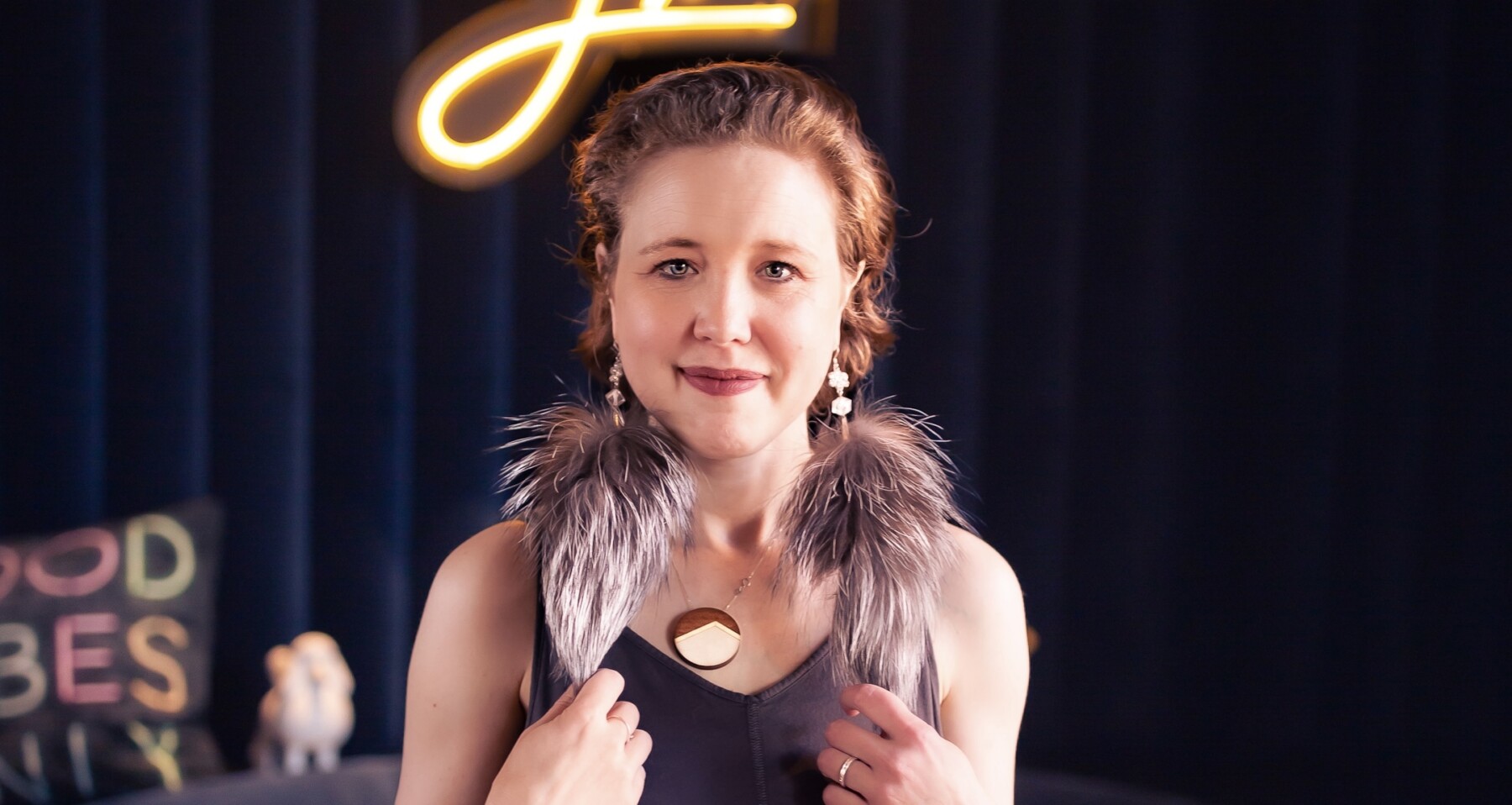 A colorful and well-composed headshot of a woman with an intriguing outfit and large, fluffy, feathered earrings, set to a black background.