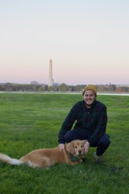 A man squats beside his dog, possibly a golden retriever, on a lush green lawn. He's wearing a yellow beanie and a flannel shirt, and both his hands are resting on his dog. The horizon is tinged with pink as the sun sets, and the Washington Monument is visible in the background.