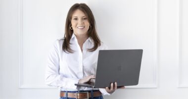 A woman in a white shirt and blue jeans stands before a white wall holding an open laptop. She's looking straight at the camera and smiling.