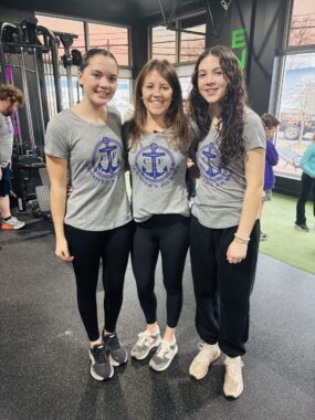 A mother stands in the center of her two daughters, all three with their arms around one another while standing and posing for a photograph. They look to be in a gym and are wearing matching outfits: black yoga pants and gray T-shirts with a blue anchor on the front. The two daughters are slightly taller than their mother.