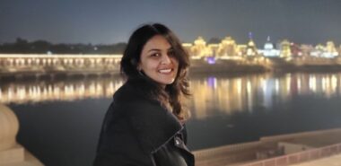 A woman poses for a photo in front of a river at nighttime. The buildings across the water are lit up and casting reflections on the river's surface.