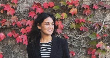 A woman poses for a photo in front of a wall covered in vines and red leaves. She's wearing a black cardigan over a striped top and is looking off to her left.