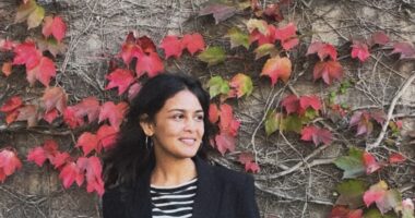 A woman poses for a photo in front of a wall covered in vines and red leaves. She's wearing a black cardigan over a striped top and is looking off to her left.