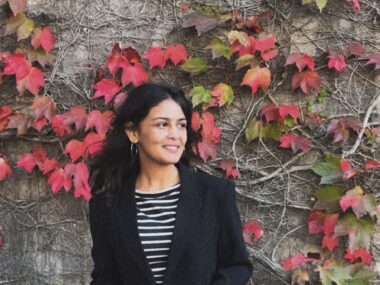 A woman poses for a photo in front of a wall covered in vines and red leaves. She's wearing a black cardigan over a striped top and is looking off to her left.