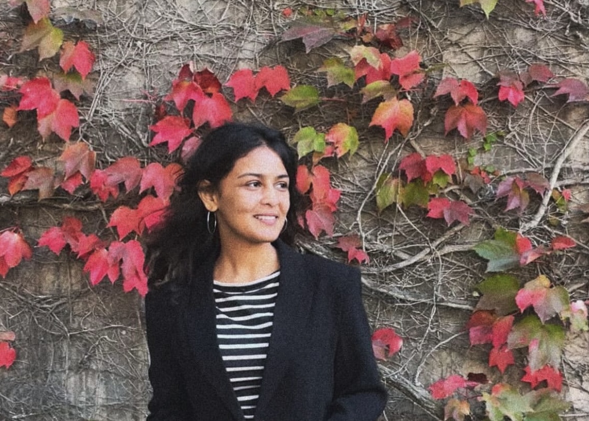 A woman poses for a photo in front of a wall covered in vines and red leaves. She's wearing a black cardigan over a striped top and is looking off to her left.