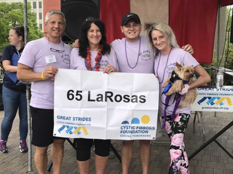 A family of four, all wearing matching purple T-shirts, poses for a photo at the Cystic Fibrosis Foundation's Great Strides event. They're holding up a sign that says "65 LaRosas."