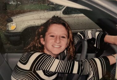 A teenage girl, about 16 or 17 years old, sits behind the wheel of a car with her arms outstretched in front of her. She's wearing a long-sleeve black-and-white striped shirt and has her head turned to the right, smiling at the person taking her photo.