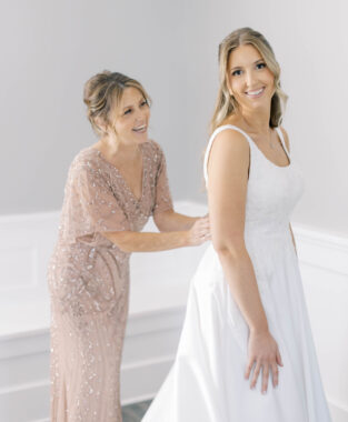 A woman stands behind another woman in a bridal gown, fastening a clasp.