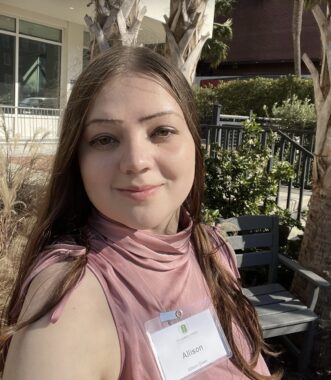 A young woman with long brown hair sits outside in the sunshine. She's wearing a pink sleeveless top, a name tag that says "Allison," and her head is turned to the right as she smiles softly at the camera.
