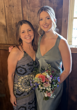 Two women, a mother and daughter, embrace and smile for a photo. The daughter holds a colorful bouquet of flowers.