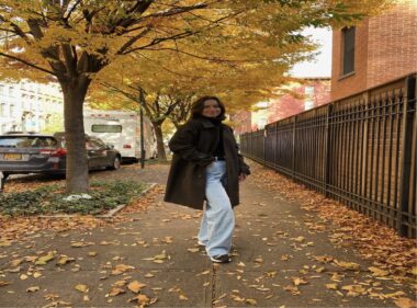 A woman wearing jeans and a long brown coat poses for a photo on a sidewalk, surrounded by trees and colorful autumn leaves.