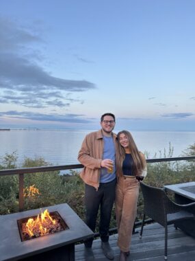 A couple stands smiling on an outdoor deck at dusk, next to a square fire pit with an open flame. The man wears a tan jacket and holds a beer; the woman has long brown hair and holds a glass of red wine. A calm body of water and soft pink-blue sky stretch out behind them.
