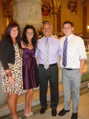 A family of four – mother, father, son, and daughter - stand side by side and smile for a photo. They are all dressed formally and are standing inside a large, dimly lit room.